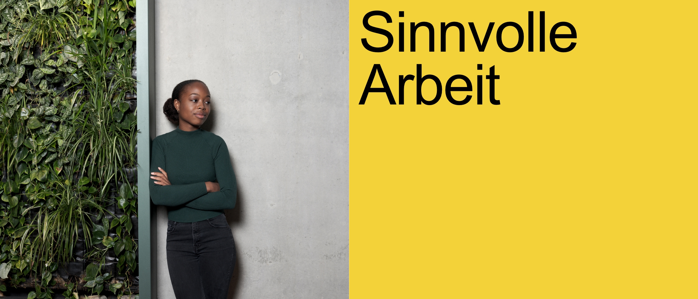 Sinnvolle Arbeit [eine moderne Frau mit dunkler Hautfarbe lehnt mit verschränkten Armen an einer Wand an. Die eine Seite der Wand ist aus Beton, rechts ist sie mit grünen Pflanzen bedeckt. Die Frau trägt einen dunkelgrünen Wollpullover und eine schwarze Jeanshose.]