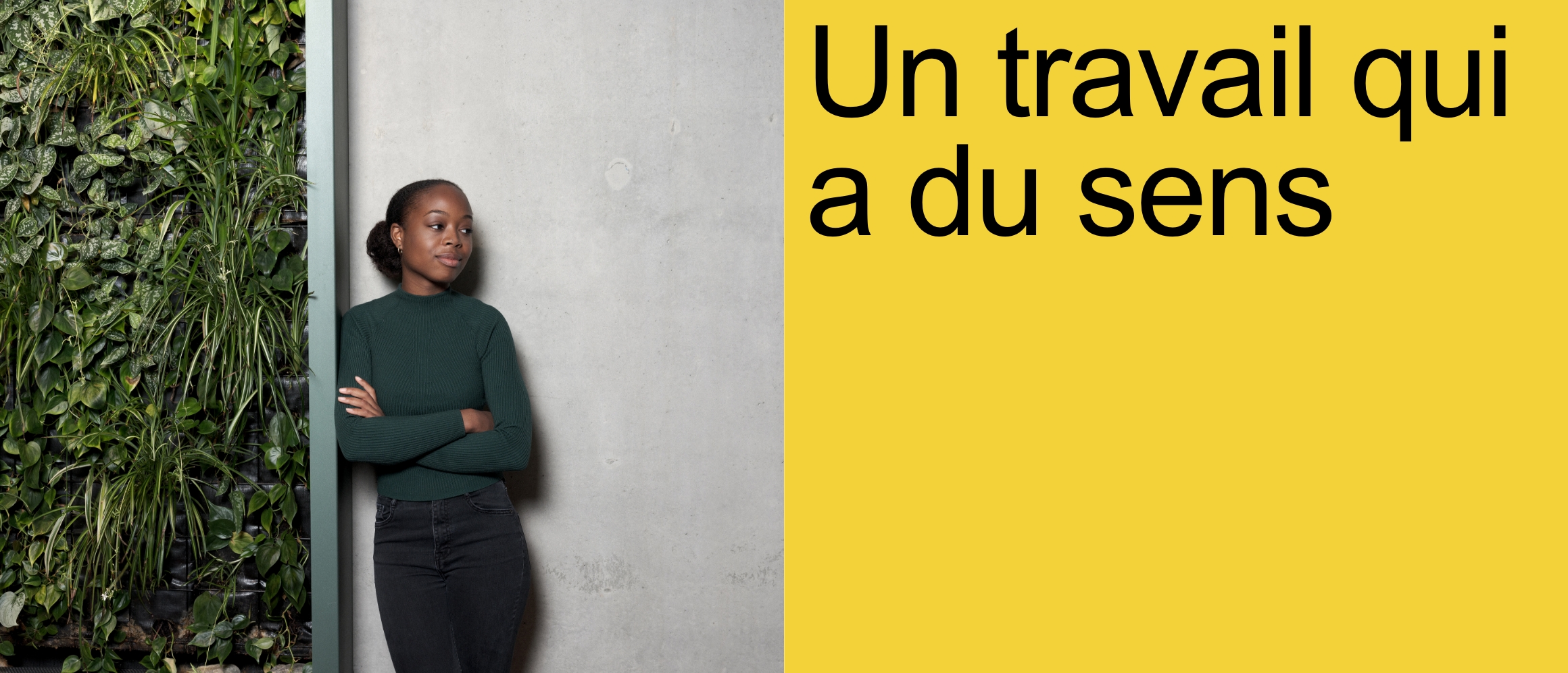 Un travail qui a du sens [une femme moderne à la peau foncée s'appuie contre un mur, les bras croisés. Un côté du mur est en béton, à droite, il est recouvert de plantes vertes. La femme porte un pull en laine vert foncé et un pantalon en jean noir.]