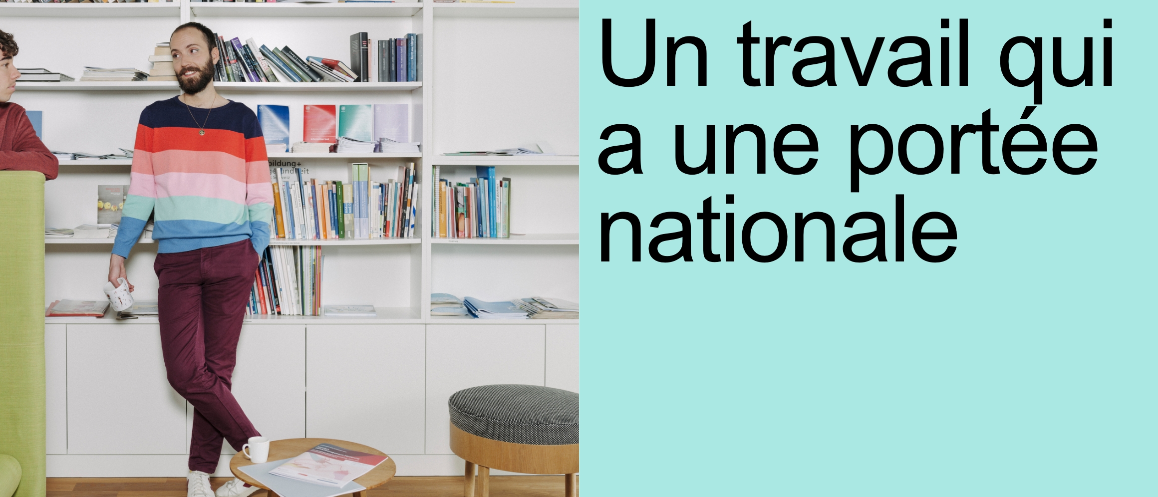 Un travail qui a une portée nationale [un jeune homme au teint clair, aux cheveux foncés courts et à la barbe coupée court se tient devant une grande bibliothèque. Il porte un pull à rayures multicolores et un pantalon rouge foncé. Il a mis sa main gauche dans la poche de son pantalon et tient dans sa main droite une tasse à café blanche avec un motif dessus. Il discute avec un collègue de travail qui se tient à sa droite.]